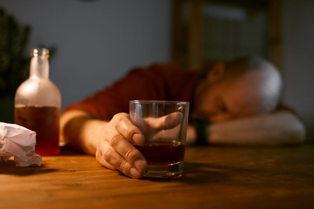 A man slumped over a table with a glass and bottle of alcohol, illustrating the struggles faced before seeking help at Huntington Beach alcohol rehab.