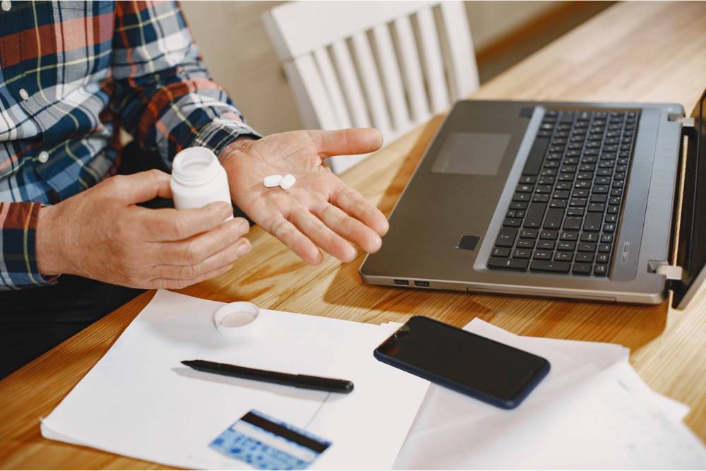 A person holding a white prescription pill bottle and pouring pills into their hand while sitting at a desk with a laptop — representing prescription drugs addiction treatment in Huntington Beach, CA.